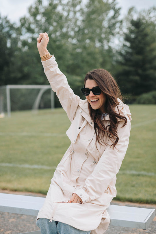 Sports Woman in a beige coat and sunglasses raising her arm outdoors on a grassy field.